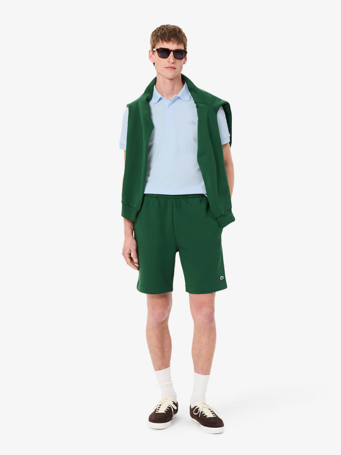 Man wearing a green sweatshirt over the shoulders, blue polo, and shorts on a white background